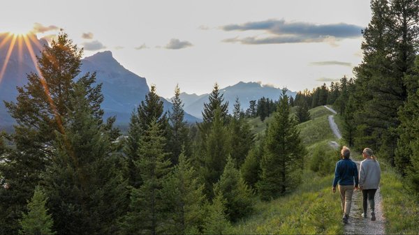 Two people walking along a trail in the mountains as the sun sets over a mountain.